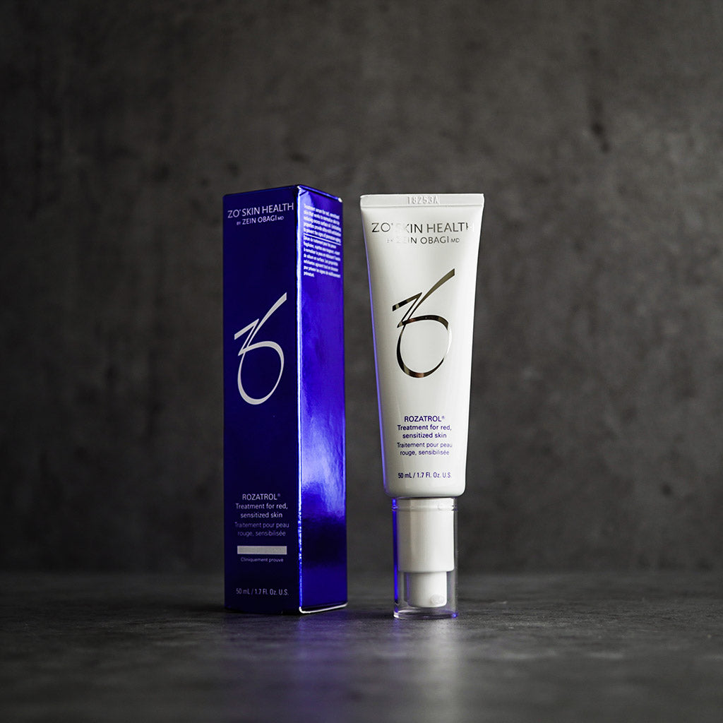 Product image of ZO Rozatrol® with its box packaging displayed against a grey concrete wall, showcasing the sleek silver and blue design, formulated to soothe redness, control oil, and support rosacea-prone skin