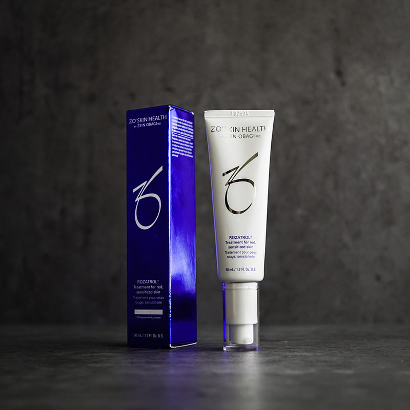 Product image of ZO Rozatrol® with its box packaging displayed against a grey concrete wall, showcasing the sleek silver and blue design, formulated to soothe redness, control oil, and support rosacea-prone skin