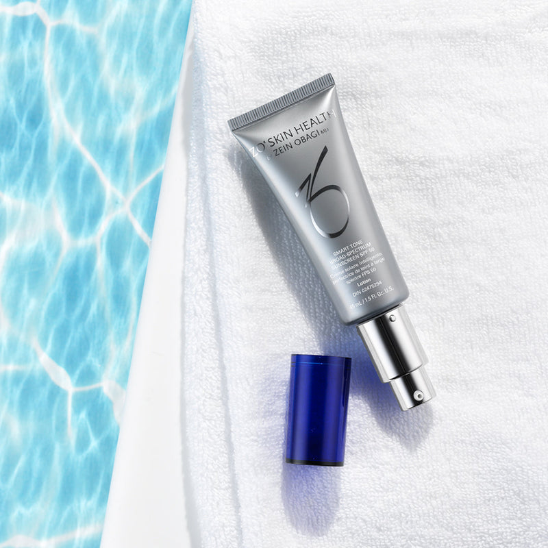 Open pump bottle image of ZO® Smart Tone Broad-Spectrum SPF 50 placed on a towel by the pool, showcasing its sleek silver and white packaging and adaptive tint formula designed to protect against UVA/UVB, blue light, and infrared rays while blending seamlessly into most skin tones