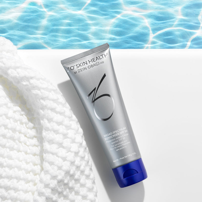 Product image of ZO® Broad-Spectrum Sunscreen SPF 50 next to a towel by the pool, featuring its sleek silver and white packaging—mineral-based and ideal for sensitive skin, offering UVA/UVB, infrared, and blue light protection with ZOX12™ antioxidants
