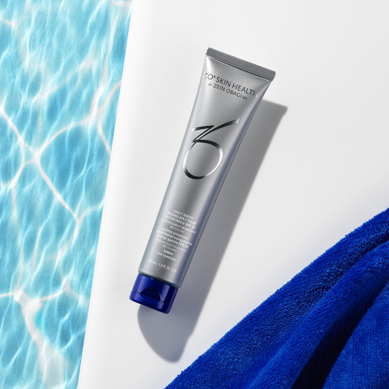 Product image of ZO® Daily Sheer Broad-Spectrum Sunscreen SPF 50 placed poolside, featuring sleek silver and white packaging—designed to provide water- and sweat-resistant protection with a sheer finish for all-day UVA/UVB, blue light, and infrared defense.